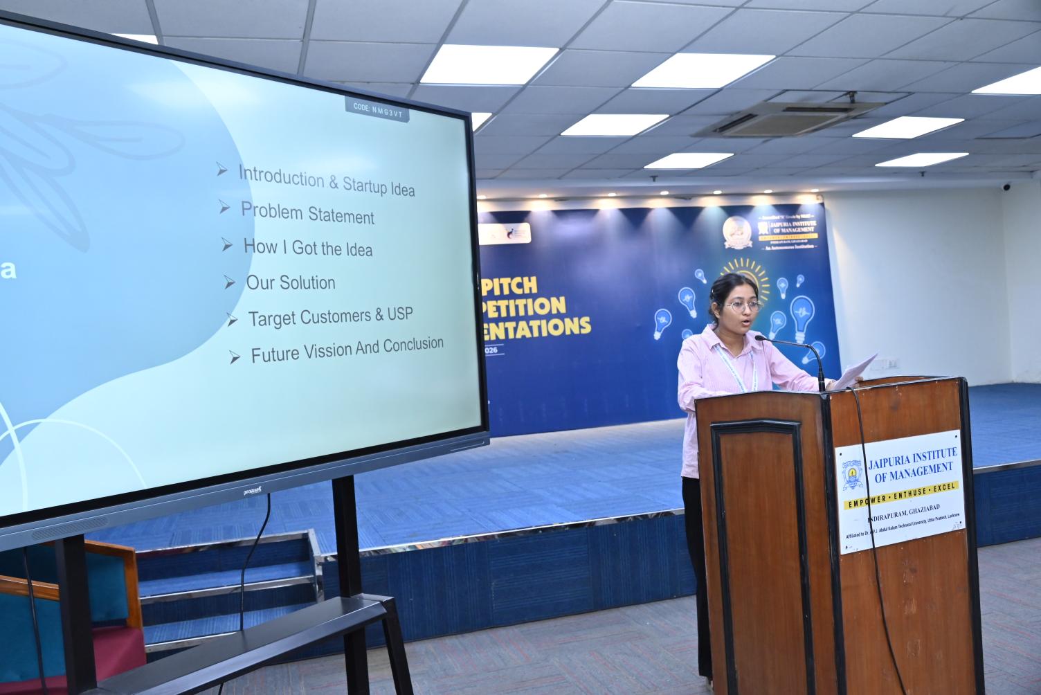 Idea Elevator Competition 2026 Turning Innovative Ideas into Entrepreneurial Opportunities (1) - Jaipuria Institute of Management, Indirapuram Ghaziabad Female presenter at a wooden podium delivering a talk; slides list bullets like Introduction & Startup Idea.