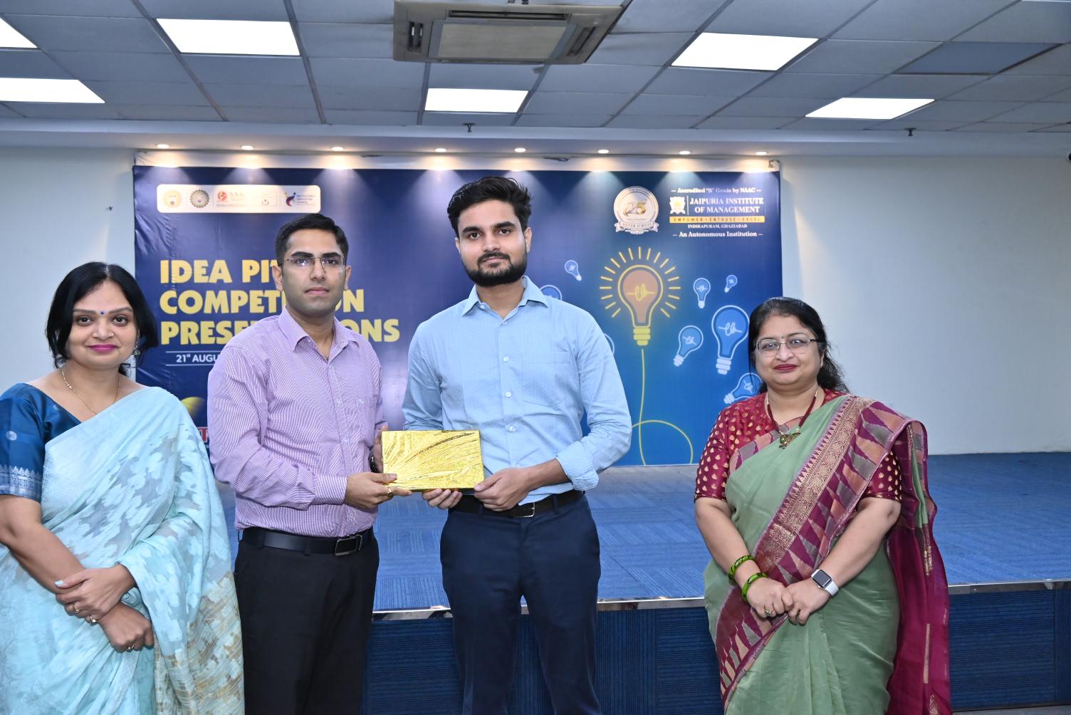 Idea Elevator Competition 2026 Turning Innovative Ideas into Entrepreneurial Opportunities (4) - Jaipuria Institute of Management, Indirapuram Ghaziabad Four people on a stage at an award ceremony; a man in a light blue shirt hands a gold-wrapped award to another man.