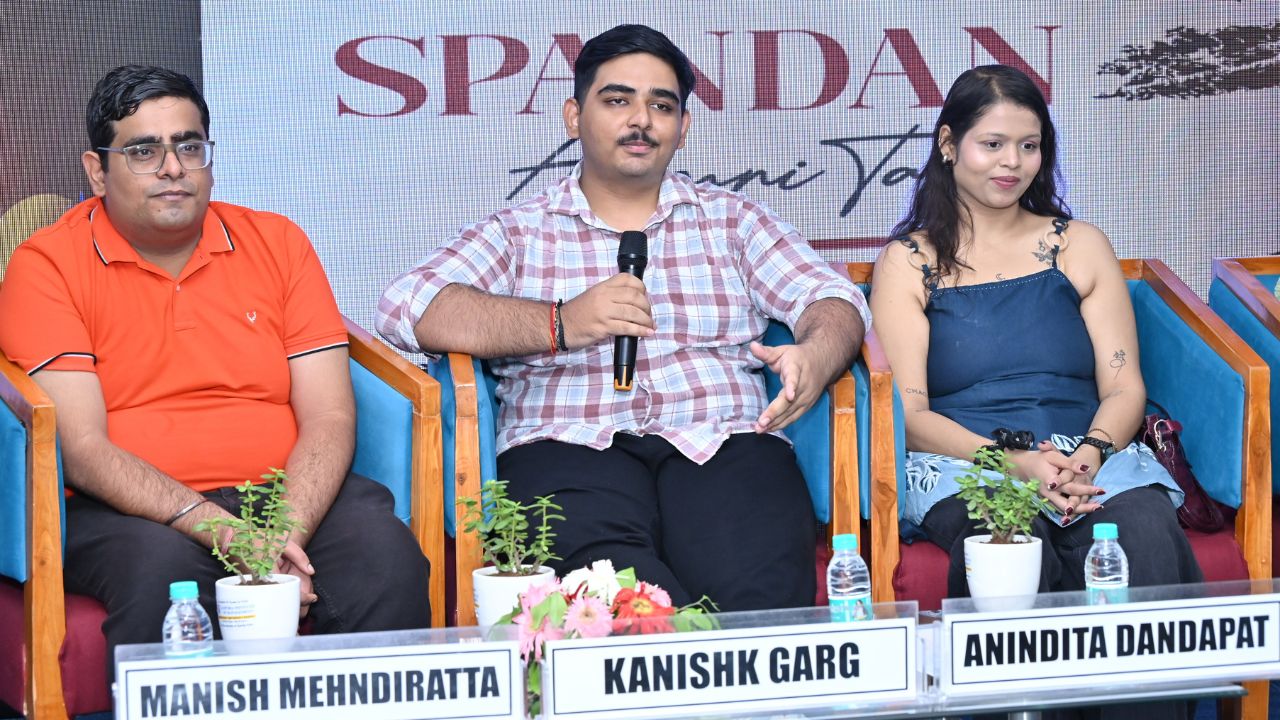 Spandan 2026 2 - Jaipuria Institute of Management, Indirapuram Ghaziabad Three panelists on a stage; center man named Kanishk Garg holds a microphone, flanked by Manish Mehndiratta and Anindita Dandapat; nameplates and small plants on the table.