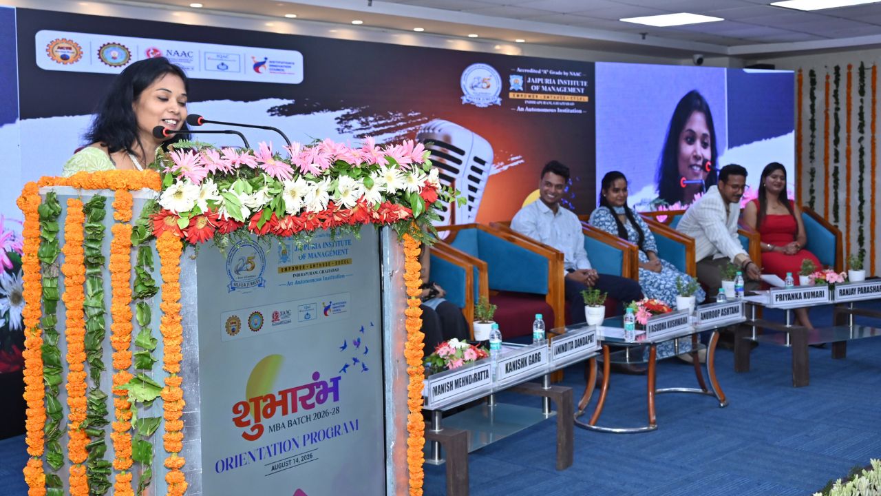 Spandan 2026 3 - Jaipuria Institute of Management, Indirapuram Ghaziabad Woman speaks at a flower-decorated podium during an orientation program; panelists sit on stage behind him/her.
