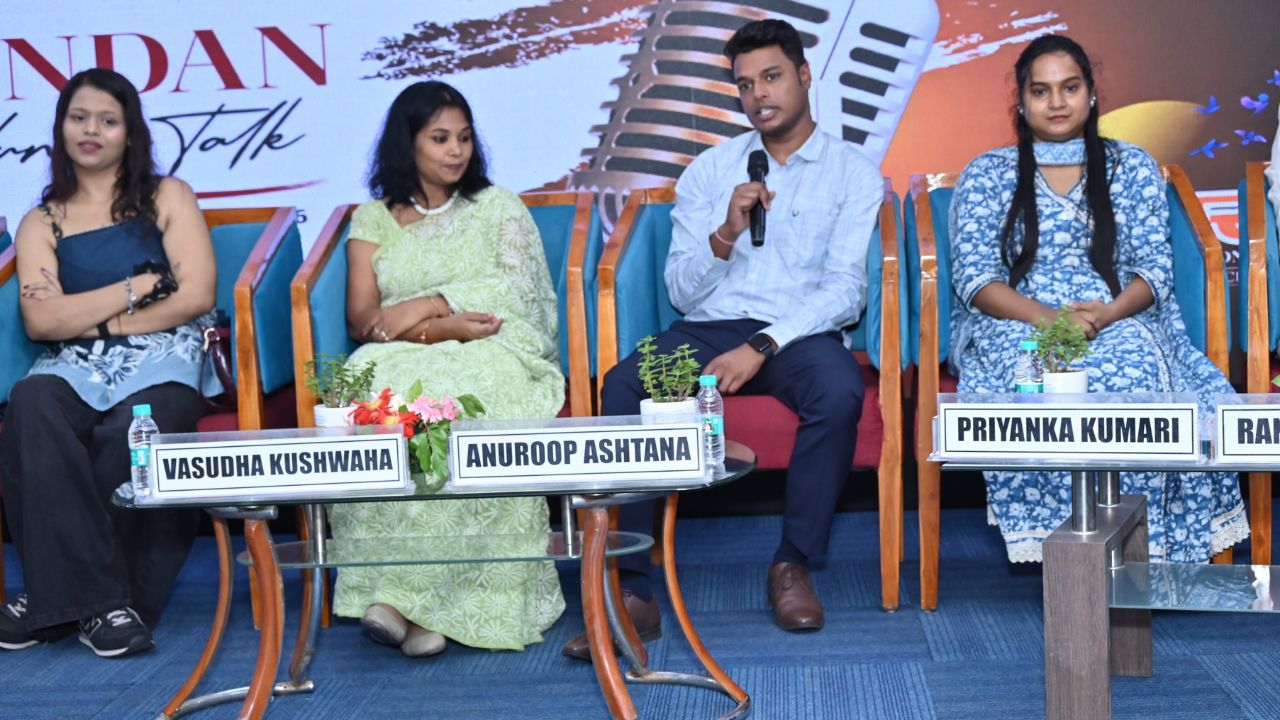 Spandan 2026 4 - Jaipuria Institute of Management, Indirapuram Ghaziabad Panel discussion with four people seated on stage; a man speaks into a microphone while three women listen, with nameplates in front reading Vasudha Kushwaha, Anuroop Ashtana, and Priyanka Kumari.