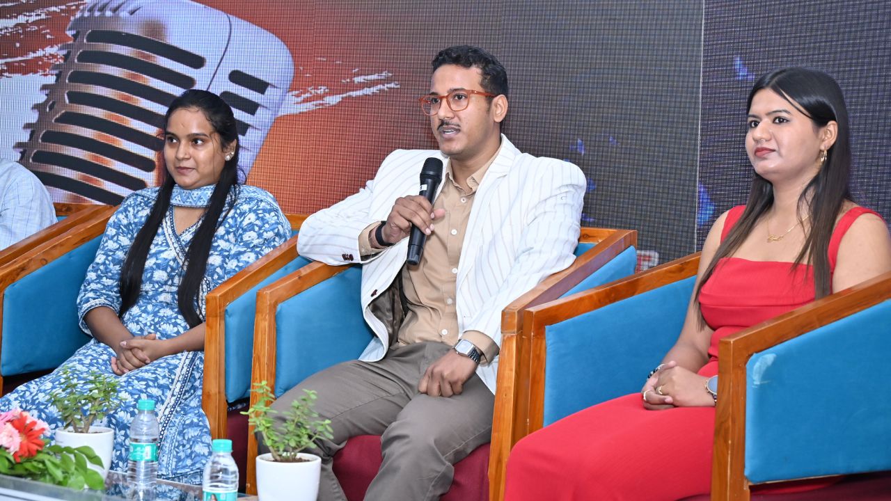 Spandan 2026 5 - Jaipuria Institute of Management, Indirapuram Ghaziabad Panel discussion with three speakers seated in blue chairs; man in center holds a microphone and speaks, flanked by two women on stage.