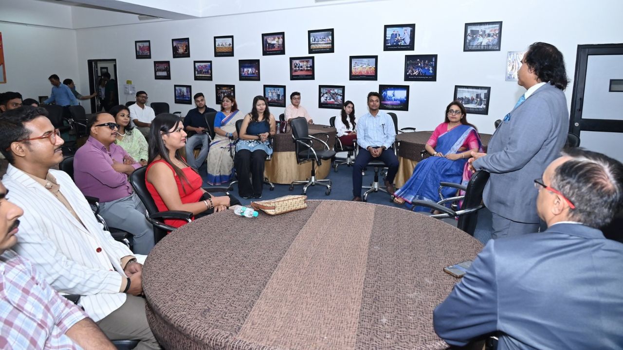 Alumni Chapter Meet NCR Chapter_ Reconnecting Legacy Celebrating Success 1 - Jaipuria Institute of Management, Indirapuram Ghaziabad A diverse group of colleagues seated in a circle around a large table, listening to a man in a suit who is speaking in a conference room with photo frames on the wall.