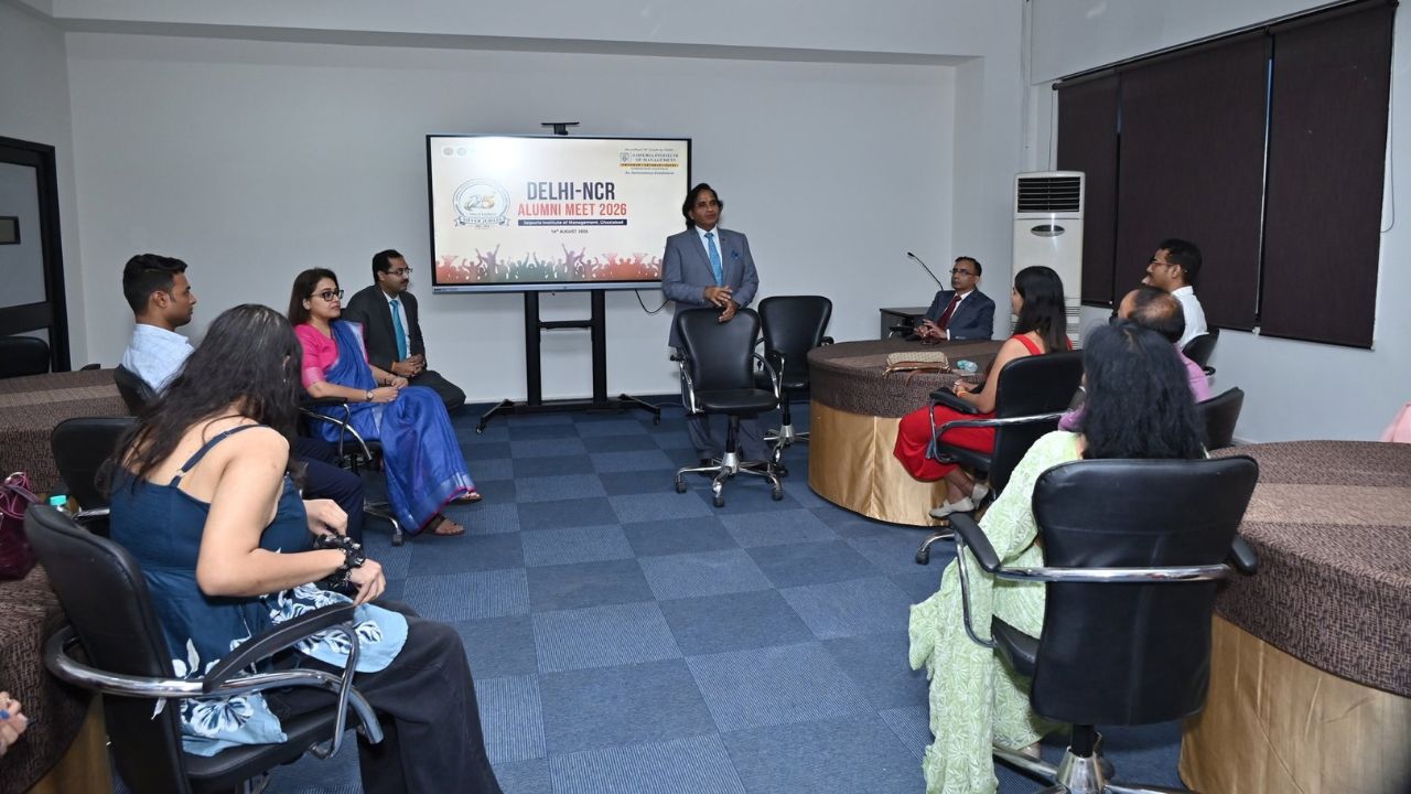 Alumni Chapter Meet NCR Chapter_ Reconnecting Legacy Celebrating Success 2 - Jaipuria Institute of Management, Indirapuram Ghaziabad A man in a suit stands and speaks to a semicircle of seated attendees in a conference room, a presentation screen behind him reads 'Delhi-NCR Alumni Meet 2026'.