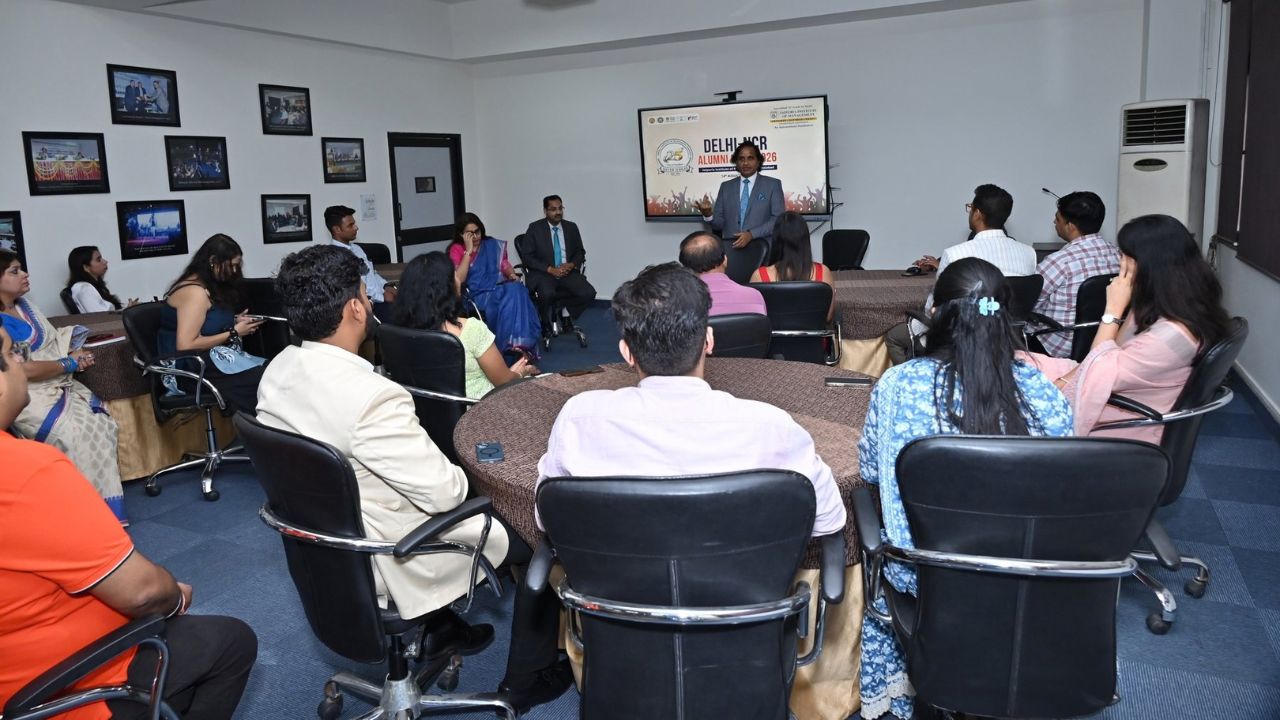 Alumni Chapter Meet NCR Chapter_ Reconnecting Legacy Celebrating Success 4 - Jaipuria Institute of Management, Indirapuram Ghaziabad A man in a suit presents at the front of a conference room as attendees sit around round tables and a screen displays a slide behind him.