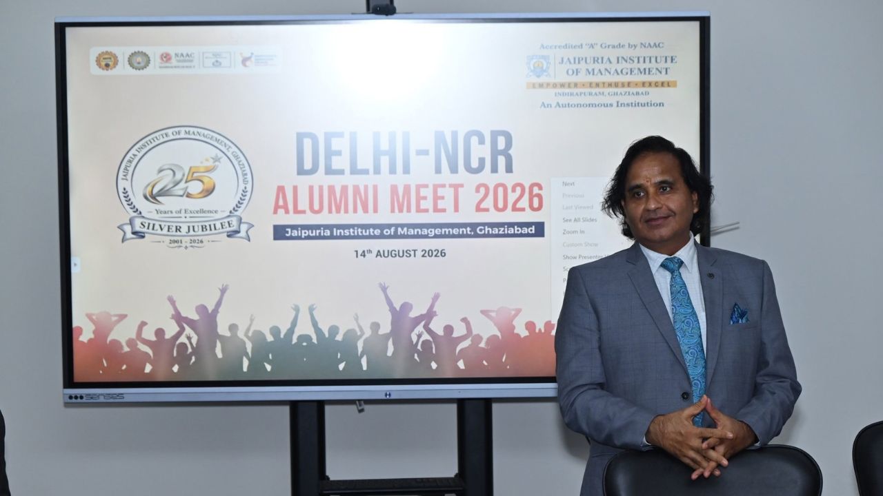 Alumni Chapter Meet NCR Chapter_ Reconnecting Legacy Celebrating Success 6 - Jaipuria Institute of Management, Indirapuram Ghaziabad Man in a gray suit and blue tie stands beside a large display screen showing the Delhi-NCR Alumni Meet 2026 banner at Jaipur Institute of Management, Ghaziabad (14 August 2026).