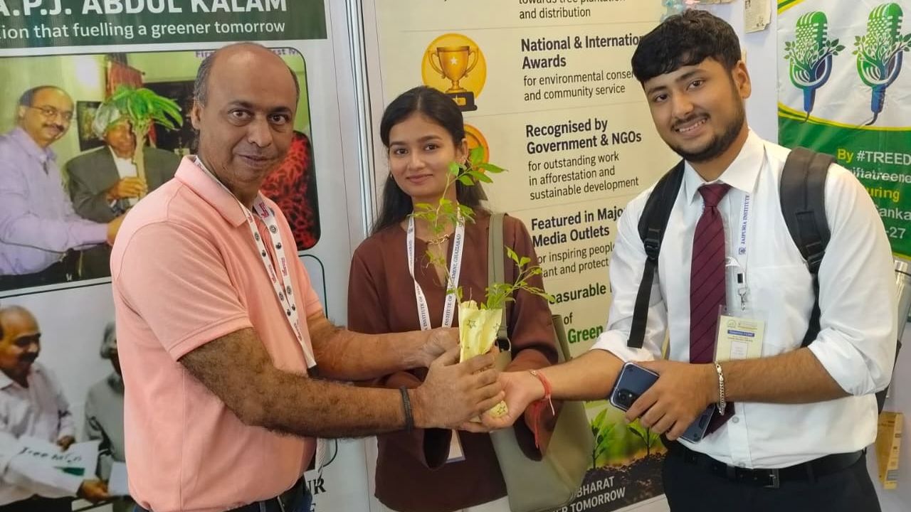 MBA Students Gain Industry Exposure at UP International Trade Show 2026 7 - Jaipuria Institute of Management, Indirapuram Ghaziabad Three people at an environmental expo exchange a small potted sapling, symbolizing a green initiative, with a man in a pink polo handing the plant to a student in a white shirt and tie while a woman stands nearby.