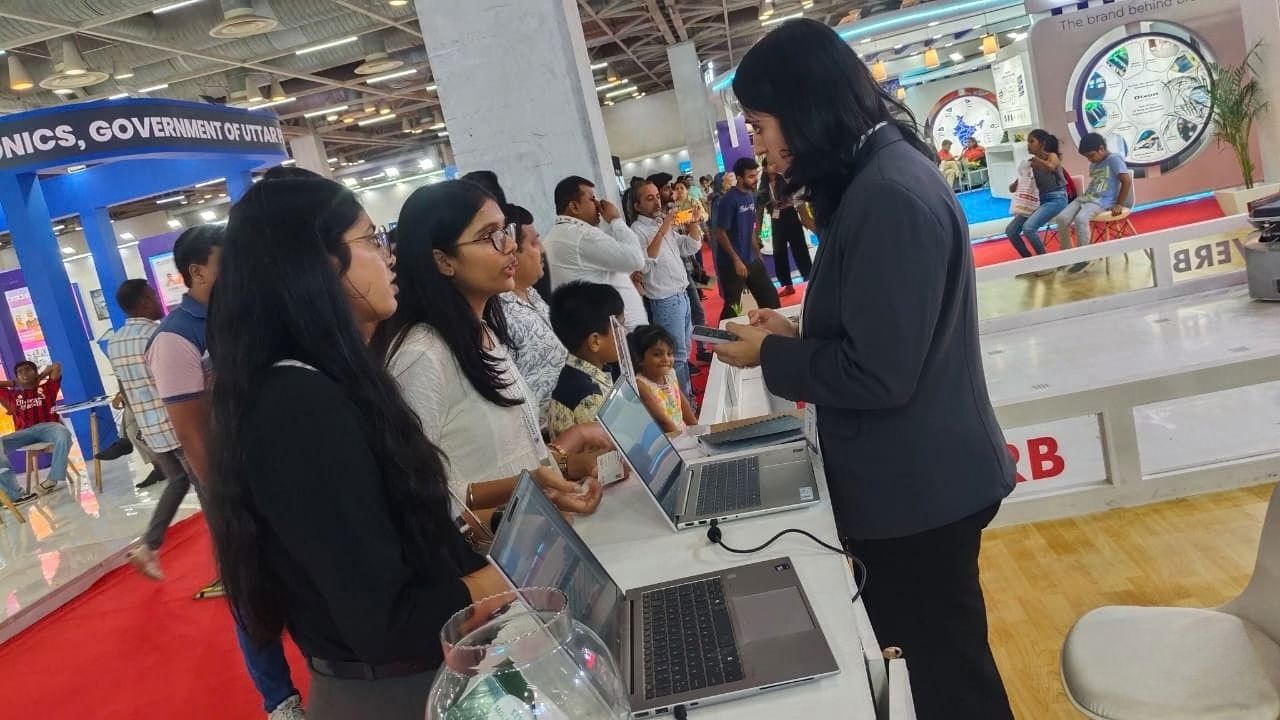 MBA Students Gain Industry Exposure at UP International Trade Show 2026 8 - Jaipuria Institute of Management, Indirapuram Ghaziabad Staff member in blazer demonstrates on a phone to attendees at a tech expo booth with laptops on the table.