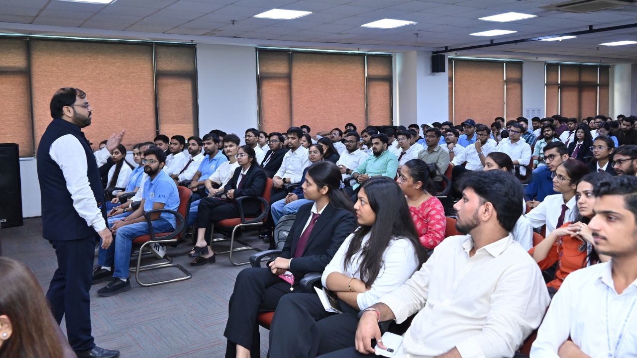Careers in Securities Markets and Role of NISM 1 - Jaipuria Institute of Management, Indirapuram Ghaziabad A man in a dark vest speaks to a large audience seated in rows of chairs in a conference room.