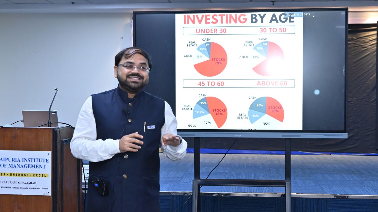 Careers in Securities Markets and Role of NISM 2 - Jaipuria Institute of Management, Indirapuram Ghaziabad Male presenter in a white shirt and navy vest speaks beside a large screen showing an 'Investing by Age' slide with four pie charts for age groups under 30, 30 to 50, 45 to 60, and above 60.