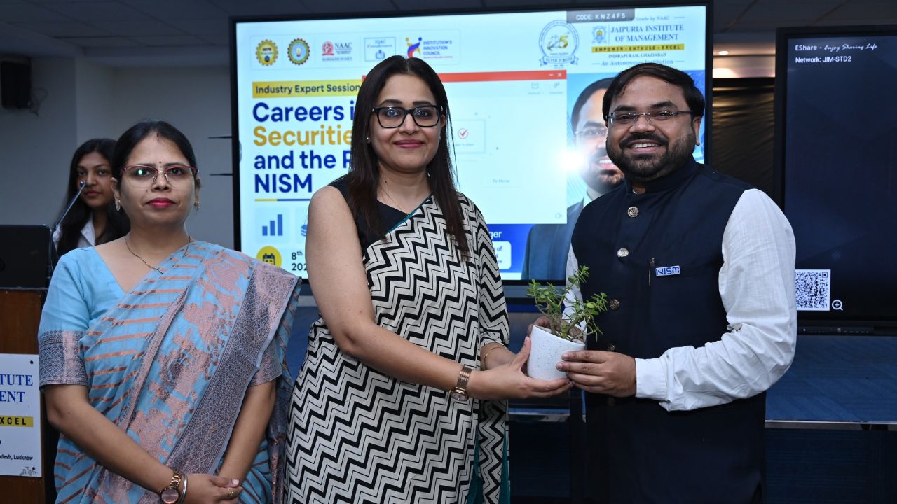 Careers in Securities Markets and Role of NISM 5 - Jaipuria Institute of Management, Indirapuram Ghaziabad Woman hands a small potted plant to a man in a dark vest during a ceremony; a presentation screen is in the background.