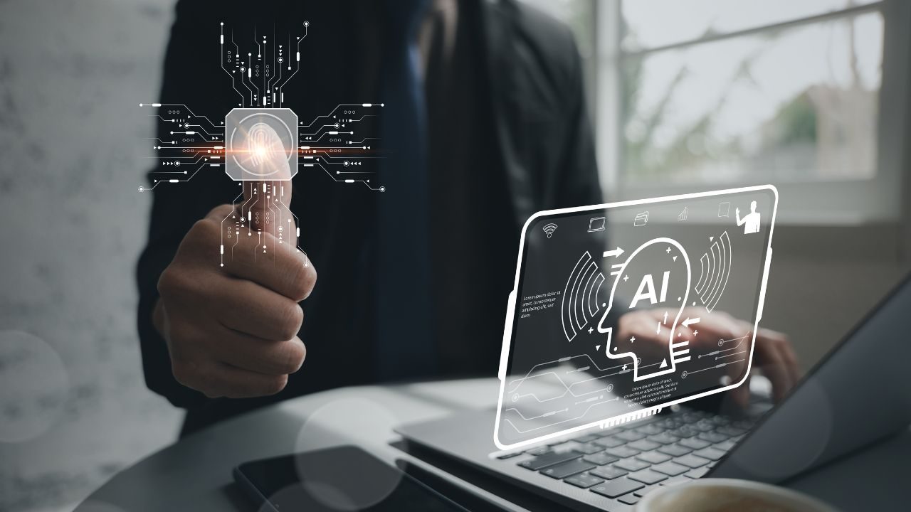Person in a dark suit holds up a glowing microchip with circuit lines projecting, while a laptop shows a holographic AI head illustration.