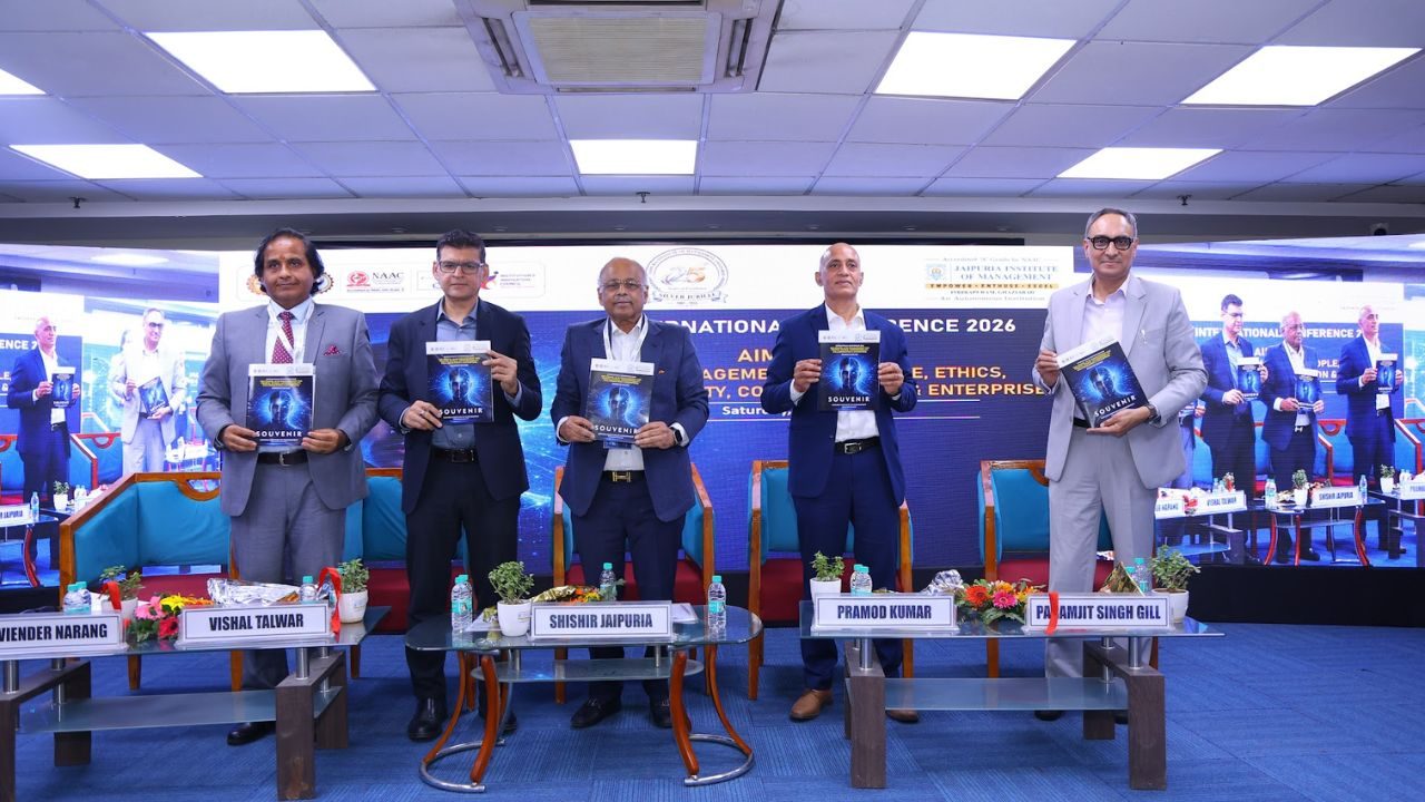 Five men in suits stand on a conference stage, each holding a blue booklet; nameplates with attendee names sit on the table in front.