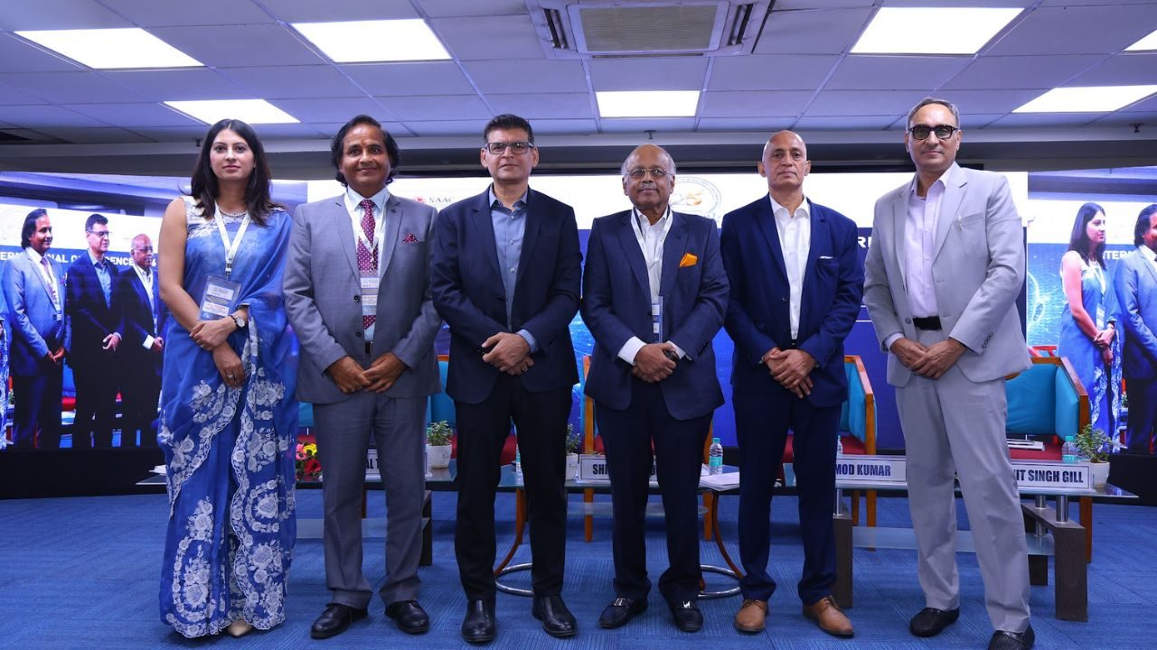 Six professionals in formal attire stand on a conference stage, posing for a group photo with a blue backdrop and screen displays behind them.