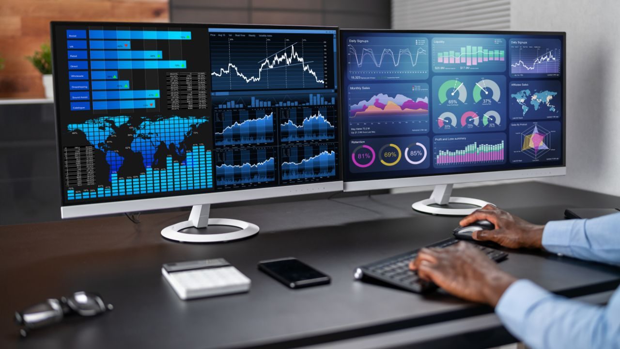 Two widescreen monitors show real-time financial dashboards with charts, maps, and graphs; hands on keyboard and mouse.