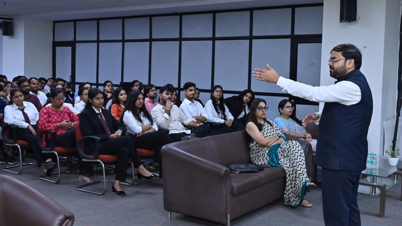 Presenter in a navy suit speaks to a classroom audience of students seated in rows.