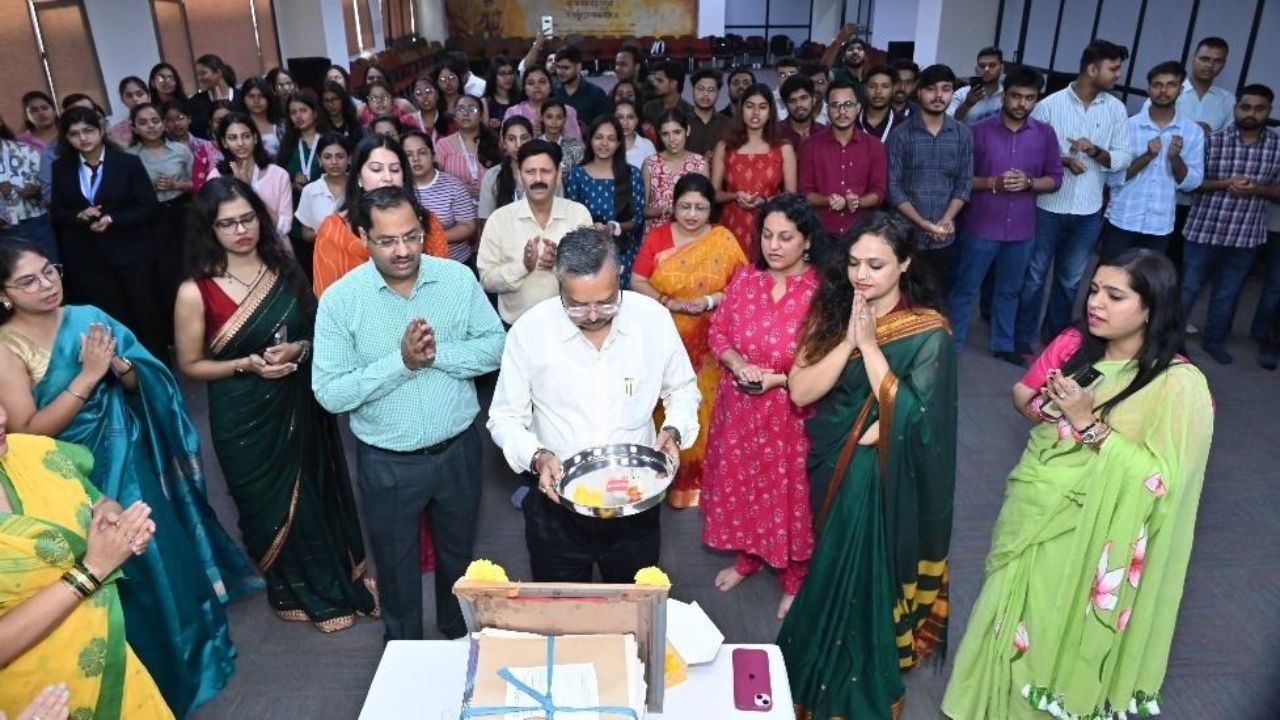 Group of coworkers gathered in a room, watching a man in a white shirt hold a tray with ceremonial items near a cake on a table in front, hinting at a celebration or ritual.