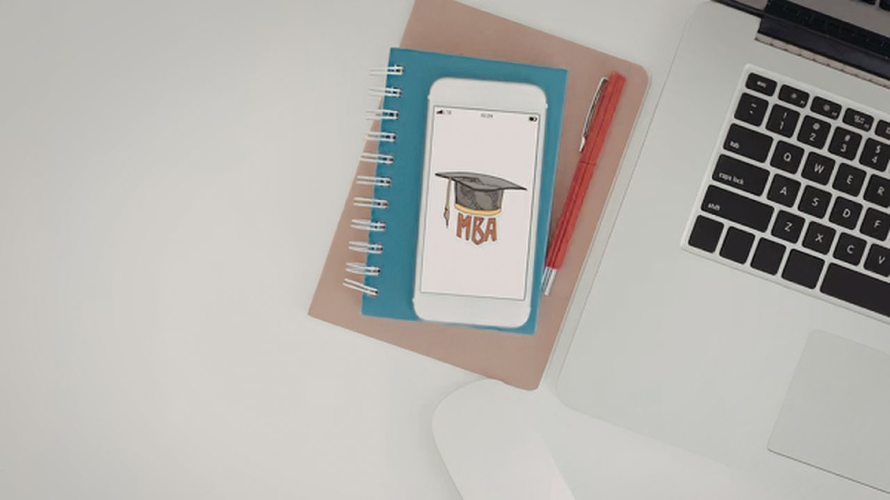 Desk setup with a laptop on the right, blue spiral notebook, a phone showing an MBA graduation cap illustration, and red pens nearby.