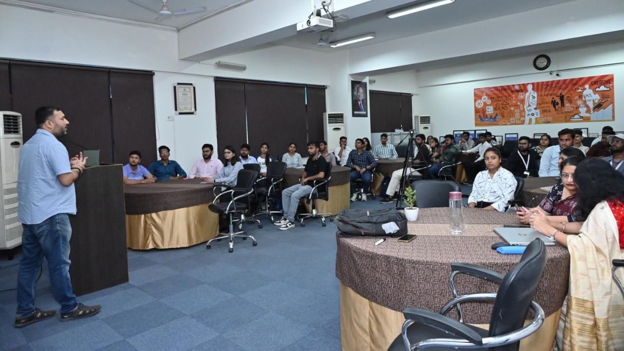 Lecturer at a podium speaks to a conference room full of seated attendees, with a projector overhead and a colorful poster on the wall behind them.