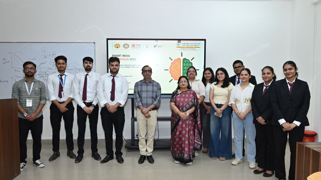 Group of students and two teachers pose for a photo in a classroom with a presentation screen in the background showing a Smart India event logo.