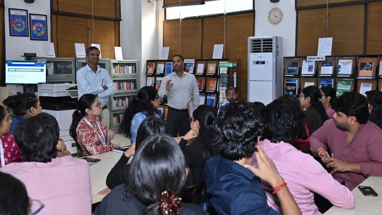 A presenter speaks to a group of students in a library, with shelves and a computer station in the background.