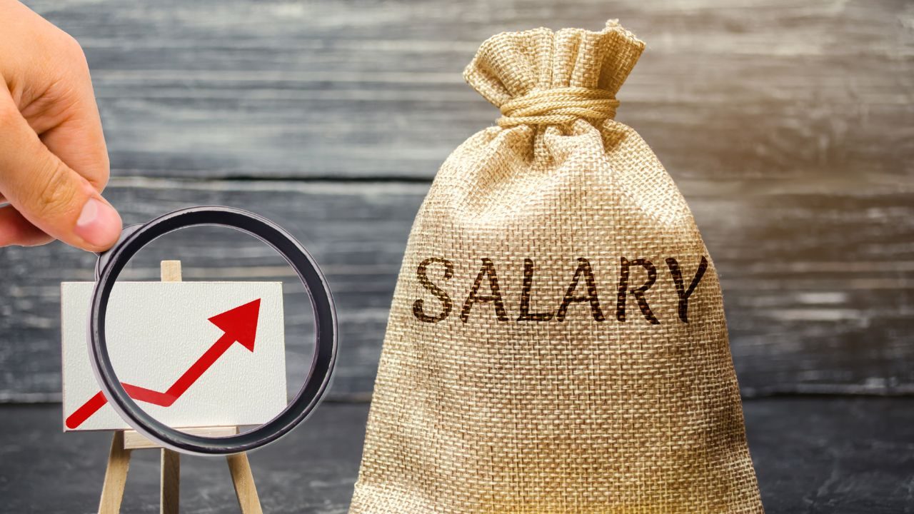 Hand holds a magnifying glass over a small chart with a rising red arrow beside a burlap sack labeled SALARY on a wooden backdrop.
