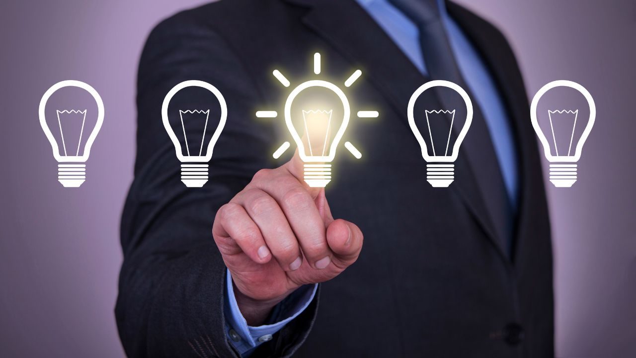 Businessperson in a suit taps a glowing light bulb among five bulbs, symbolizing a new idea or innovation.