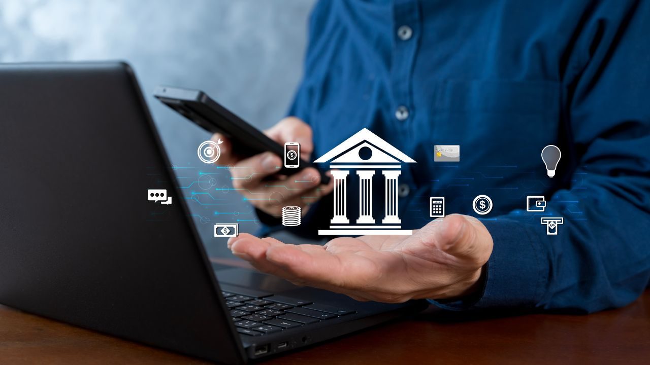 Hands above a laptop with floating fintech icons around a central bank building symbol, illustrating digital banking concept.