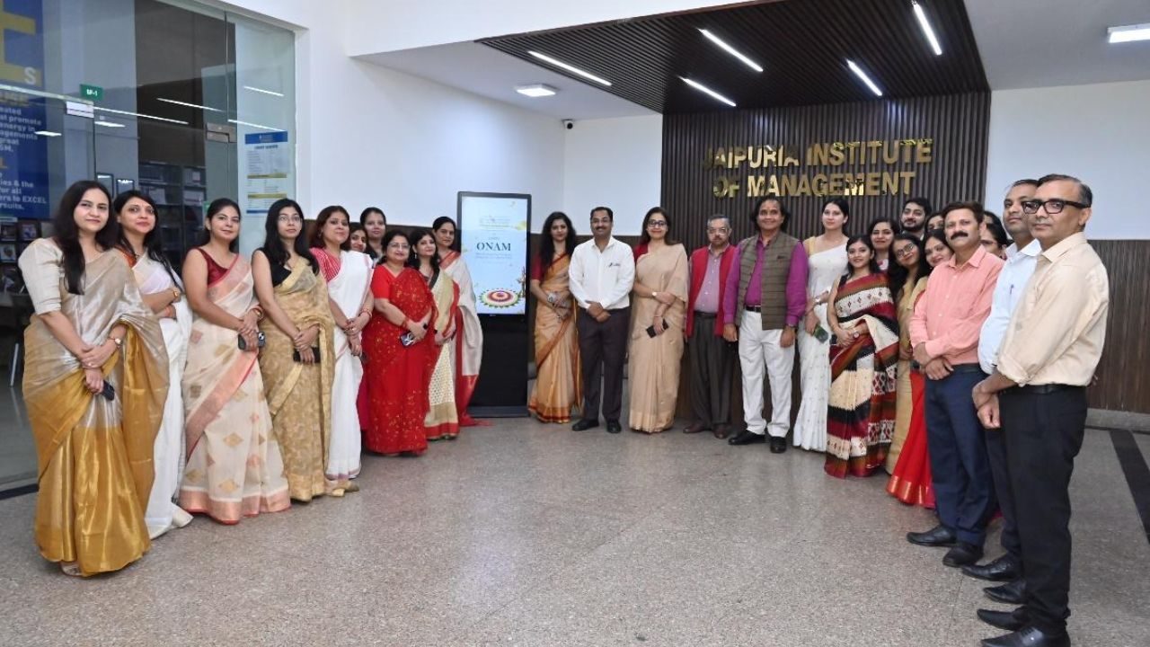 Group of men and women in formal attire posing in a university lobby in front of a wooden panel sign that reads 'AIPURIA INSTITUTE OF MANAGEMENT'.