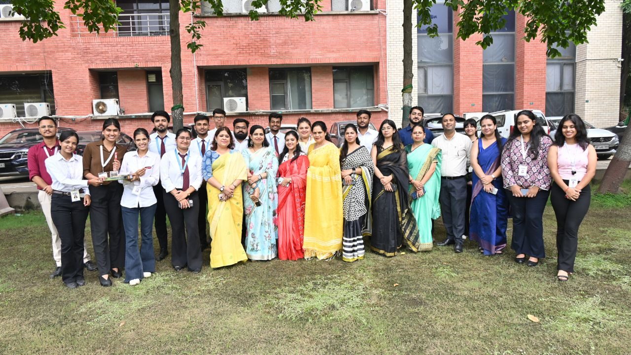 Group of men and women in formal attire posing outdoors on a grassy area in front of a red-brick building.