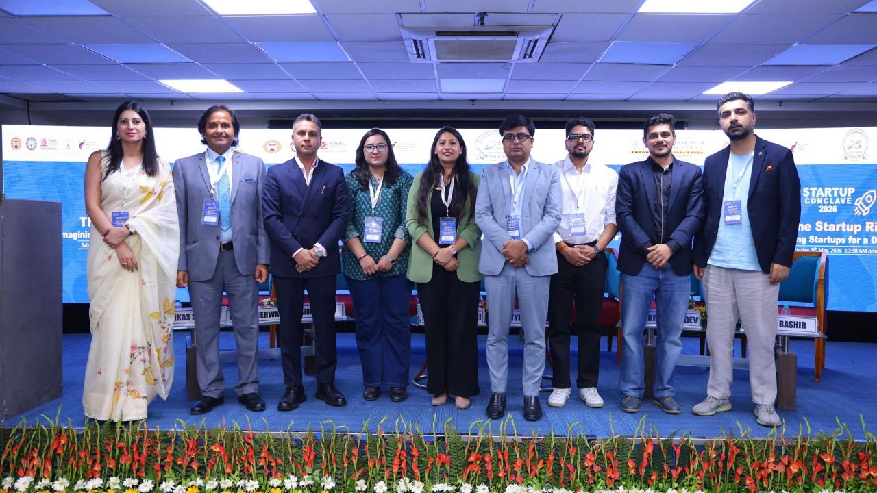Group of ten professionals on a stage at a startup conclave, posing for a photo with badges visible under bright conference lights.