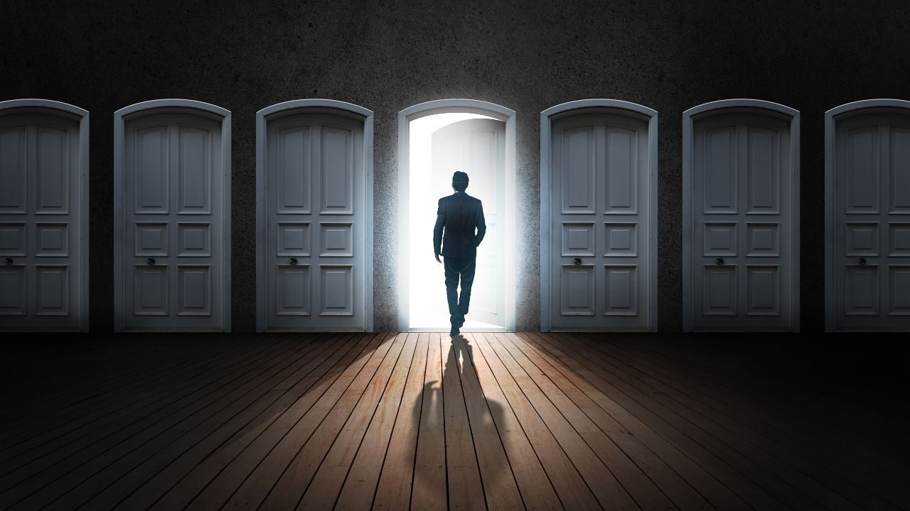 Silhouette of a man in a suit walking through a bright open door in a row of closed doors, casting a long shadow.
