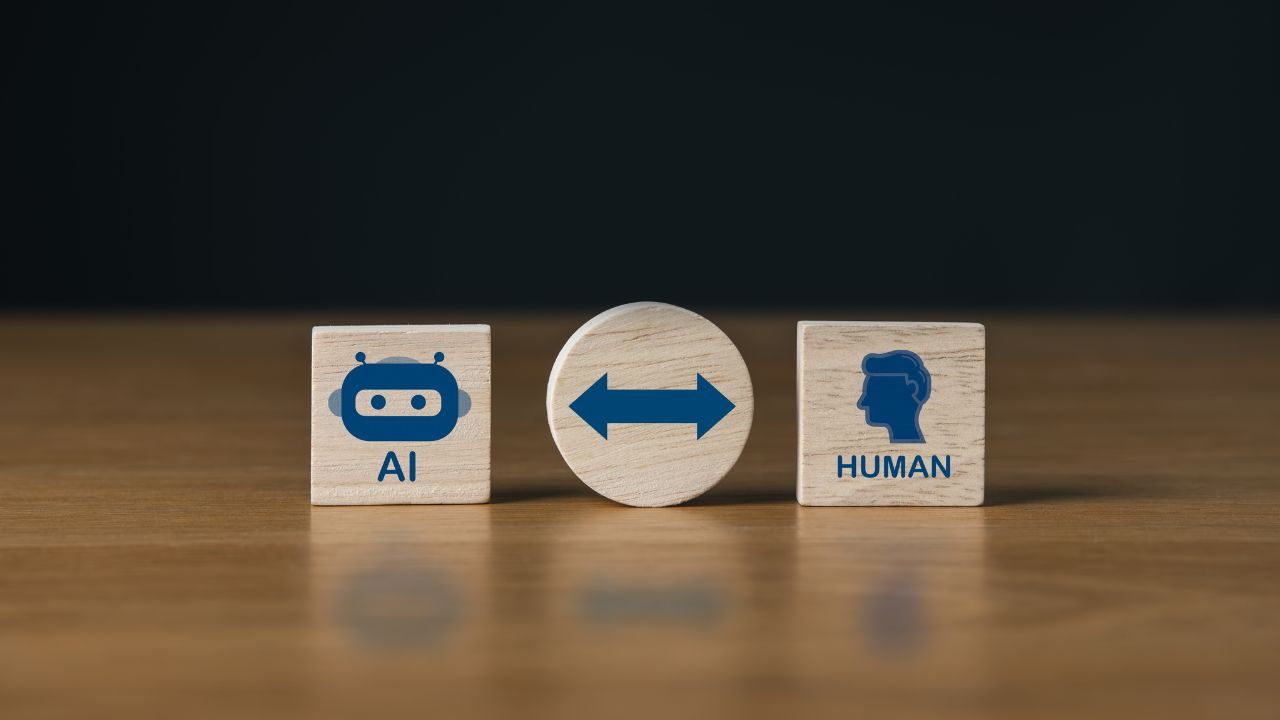 Wooden blocks show AI with a robot icon, a central double-headed arrow, and a HUMAN block with a head silhouette—illustrating human–AI collaboration.